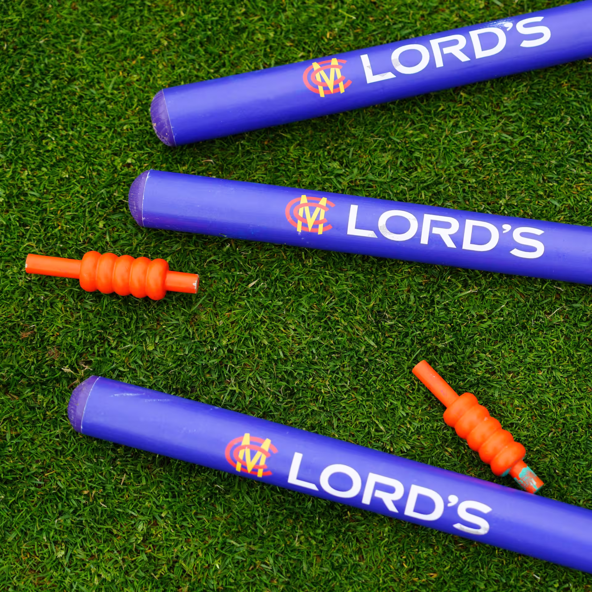Close-up of two Lord's MCC-branded blue cricket stumps and orange bails lying on green grass