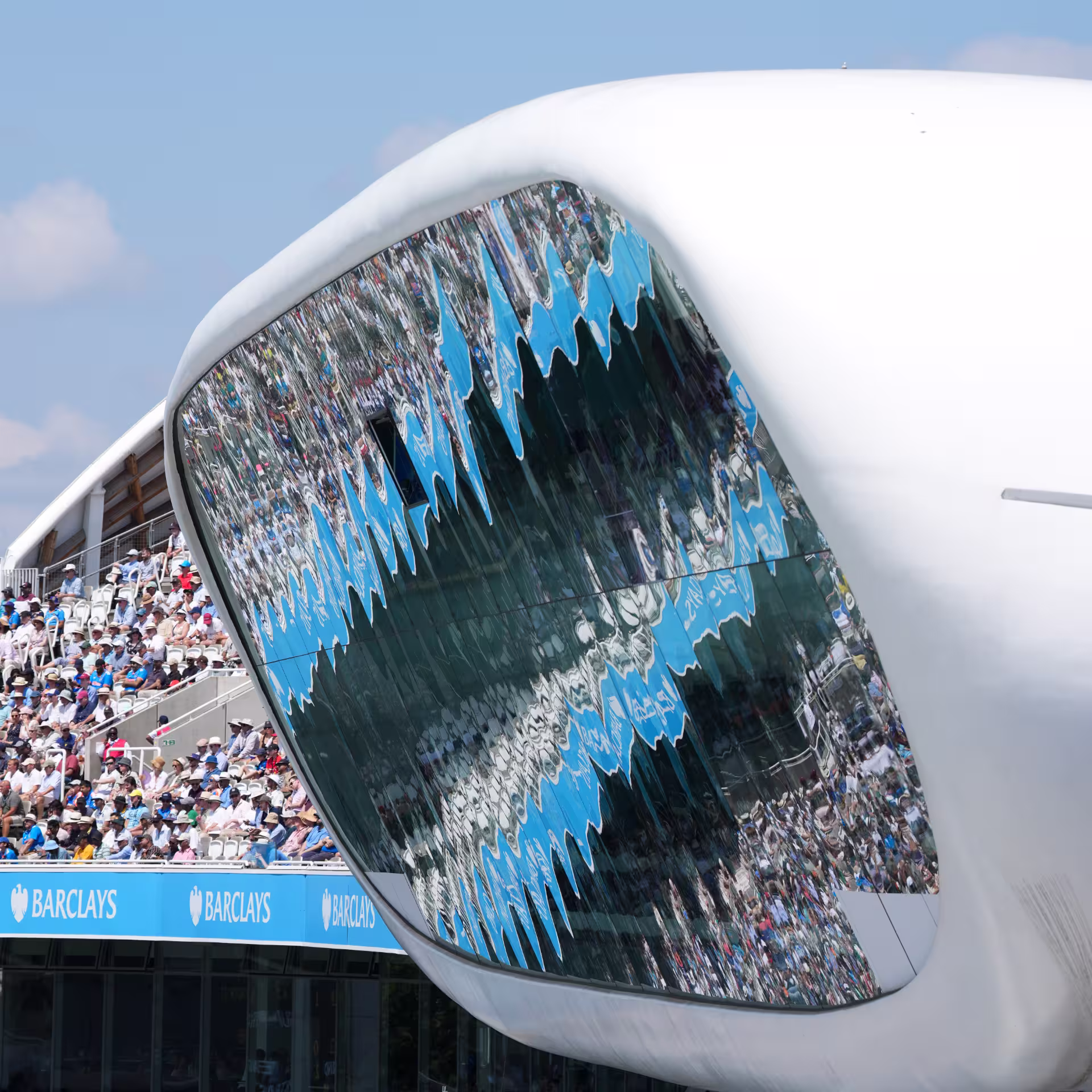 Close-up of the Lord's Media Centre with its reflective glass facade mirroring the crowd, Barclays advertising boards below, and packed stands on a sunny match day