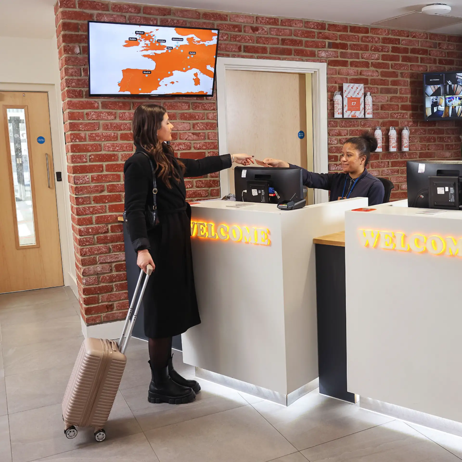 A woman in a black coat stands at a modern white hotel reception desk with a "Welcome" sign, handing her ID or card to the receptionist.