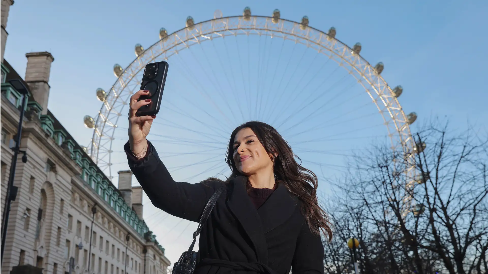 A low-angle shot of a woman taking a selfie with the London Eye in the background against a clear blue sky.
