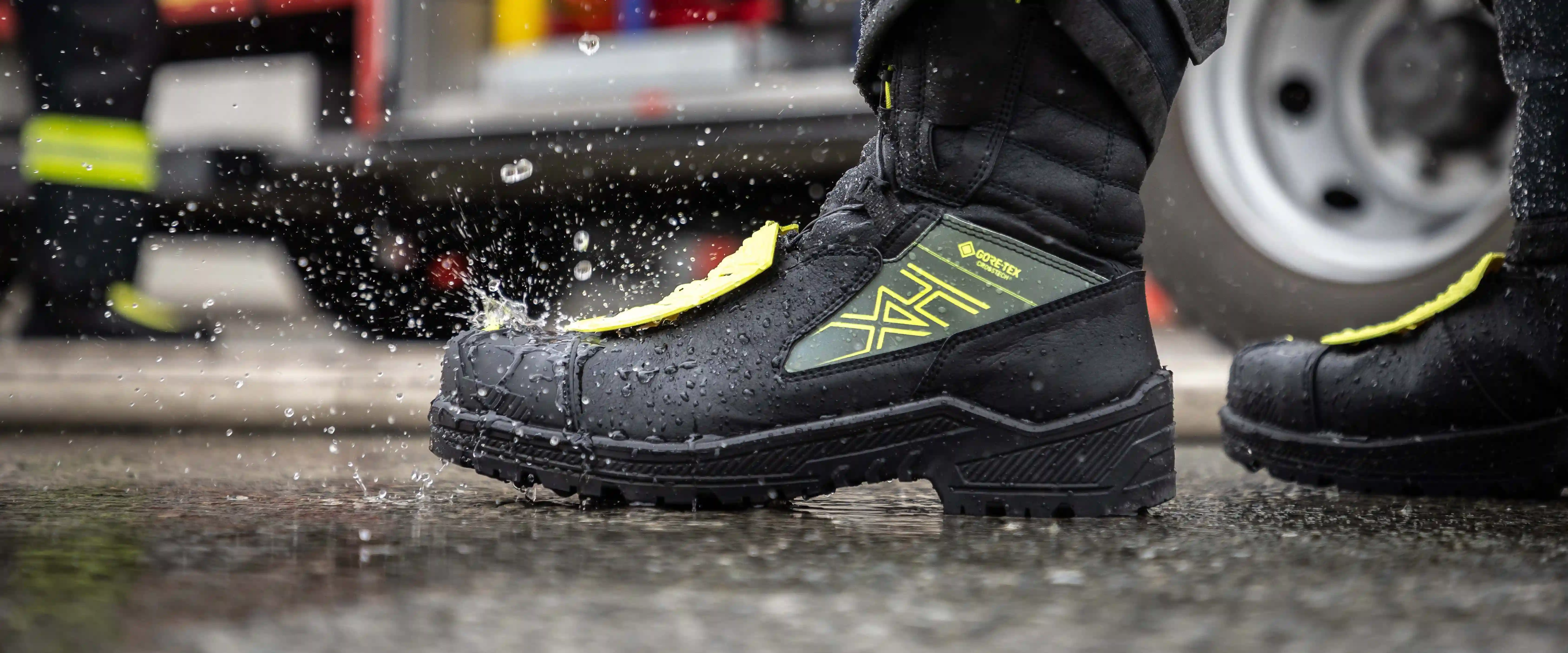 An extreme close-up, side-profile shot of a rugged HAIX fire fighter boot stepping into a rain-slicked puddle, capturing the moment of impact with a dramatic splash of water droplets. The boot is primarily black with a tactical Gore-Tex panel in olive green featuring the distinctive yellow HAIX logo, and vibrant neon yellow protective elements on the laces and heel. The background is softly blurred, showing the reflective wet pavement and the wheel of a fire truck, emphasizing the boot's durability and waterproof performance in demanding emergency environments.
