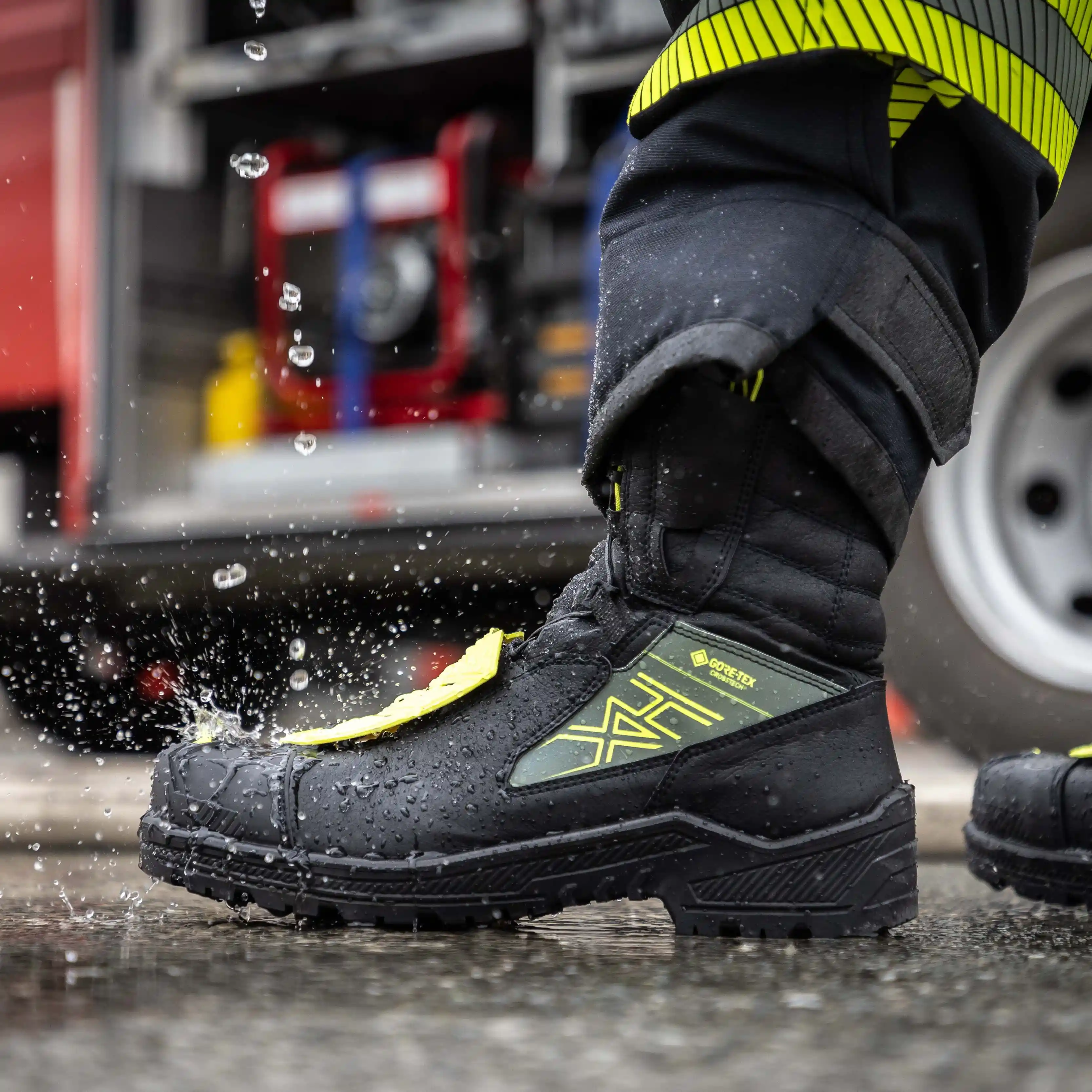 An extreme close-up, side-profile shot of a rugged HAIX fire fighter boot stepping into a rain-slicked puddle, capturing the moment of impact with a dramatic splash of water droplets. The boot is primarily black with a tactical Gore-Tex panel in olive green featuring the distinctive yellow HAIX logo, and vibrant neon yellow protective elements on the laces and heel. The background is softly blurred, showing the reflective wet pavement and the wheel of a fire truck, emphasizing the boot's durability and waterproof performance in demanding emergency environments.
