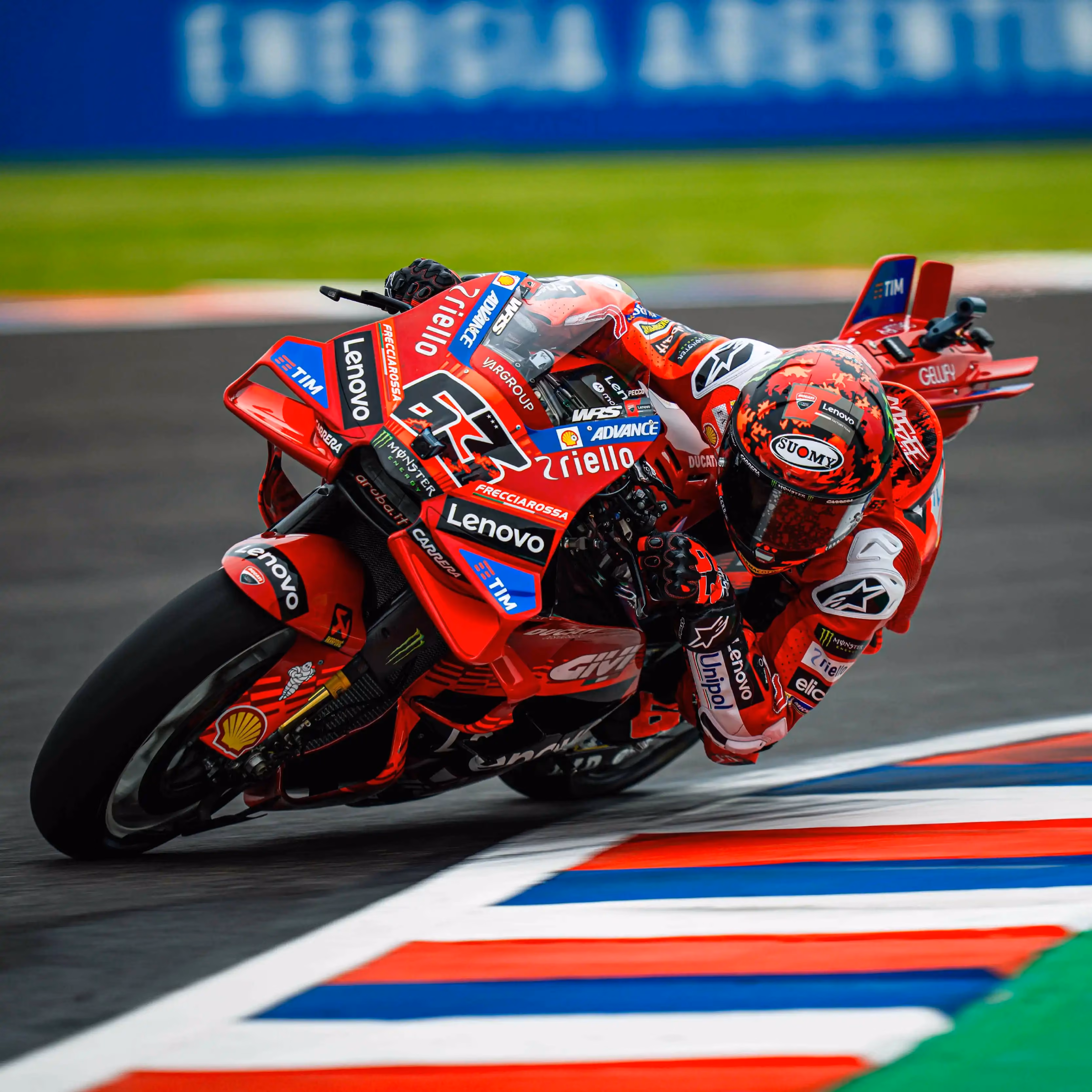 A professional motorcycle racer wearing a red Lenovo-branded suit and a red patterned Suomy helmet leans deeply into a sharp turn on a Ducati racing motorcycle. The bike is primarily red with prominent logos for Lenovo, Shell, and Riello, and is captured in a dynamic action shot as it crosses over a colorful red, white, and blue track curb. The background shows a blurred racetrack under bright daylight, emphasizing the high speed of the MotoGP session.
