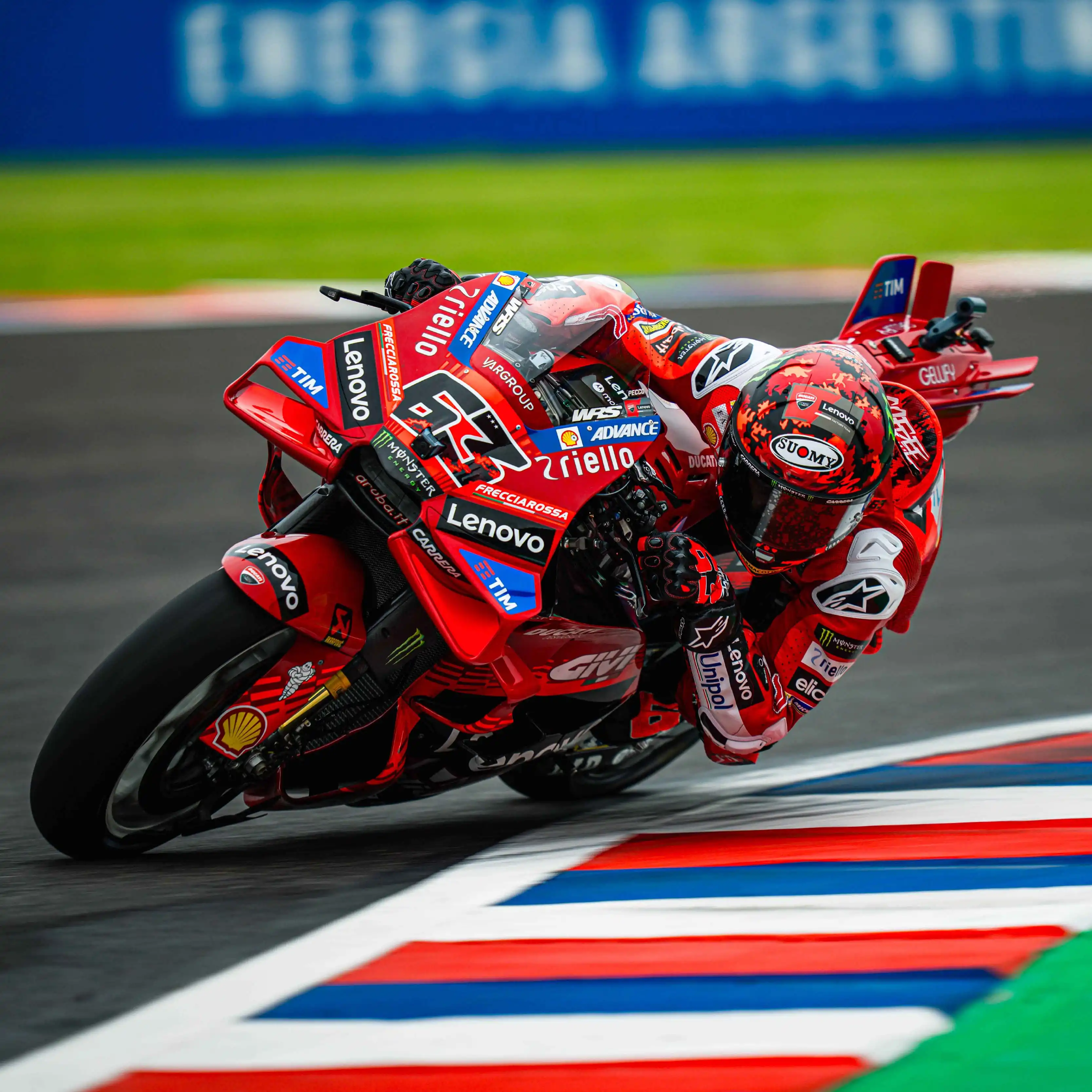 A professional motorcycle racer wearing a red Lenovo-branded suit and a red patterned Suomy helmet leans deeply into a sharp turn on a Ducati racing motorcycle. The bike is primarily red with prominent logos for Lenovo, Shell, and Riello, and is captured in a dynamic action shot as it crosses over a colorful red, white, and blue track curb. The background shows a blurred racetrack under bright daylight, emphasizing the high speed of the MotoGP session.