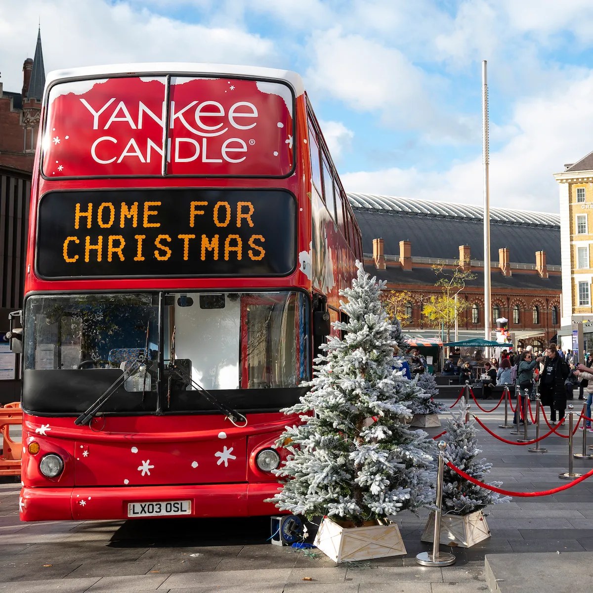 Front view of a red Yankee Candle double-decker bus with a glowing destination sign reading “Home for Christmas,” flanked by snow-covered Christmas trees. Red rope barriers mark a queue leading to a red welcome booth, and King’s Cross station and the Great Northern Hotel are in the background.