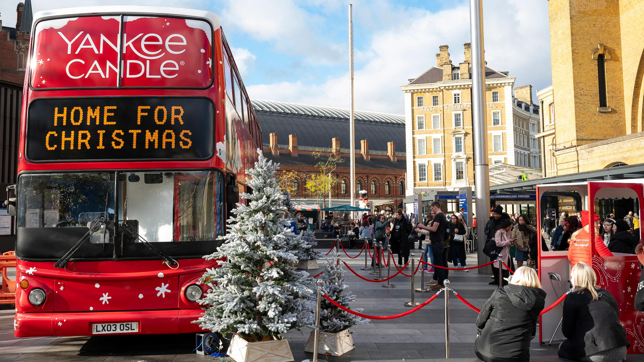 Front view of a red Yankee Candle double-decker bus with a glowing destination sign reading “Home for Christmas,” flanked by snow-covered Christmas trees. Red rope barriers mark a queue leading to a red welcome booth, and King’s Cross station and the Great Northern Hotel are in the background.