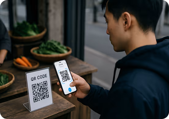 Man scanning a QR code with a smartphone at an outdoor market stall displaying fresh vegetables.