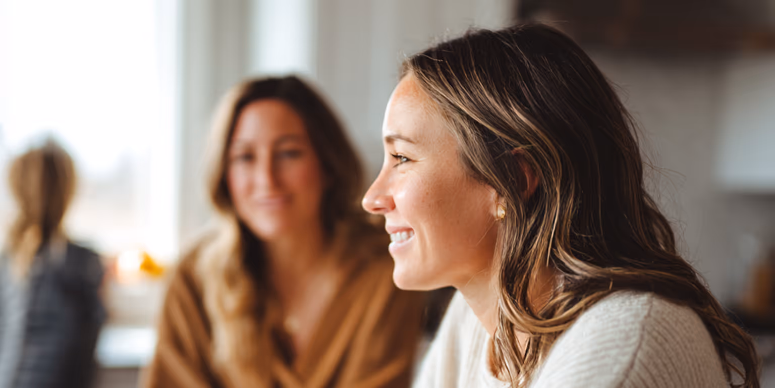 Close-up of a smiling woman with light brown hair wearing a white sweater, with another woman blurred in the background.