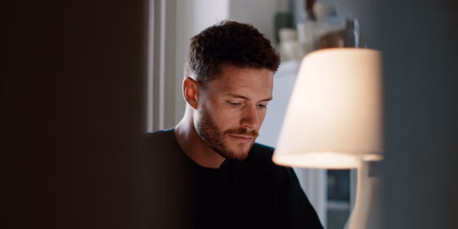 Man with short hair and beard looking downward beside a lit table lamp indoors.
