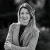 Smiling woman with long hair wearing a jacket and standing outdoors with folded arms.