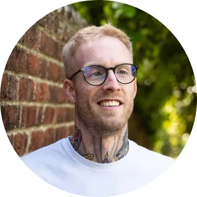 Young man with glasses and neck tattoos smiling outdoors against a brick wall.
