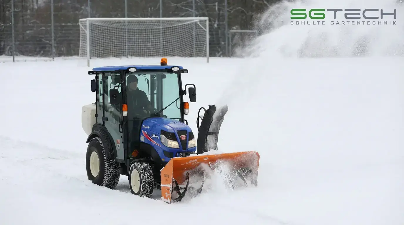 Blauer ISEKI Kompakttraktor mit orangefarbener Schneefräse räumt tiefen Schnee auf einem Fußballfeld. Eine Person sitzt in der Kabine.