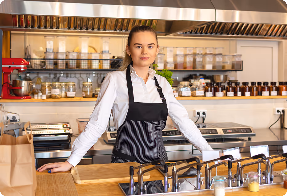Female barista wearing a white shirt and black apron standing behind the counter in a modern café with jars of ingredients and coffee-making equipment behind her.
