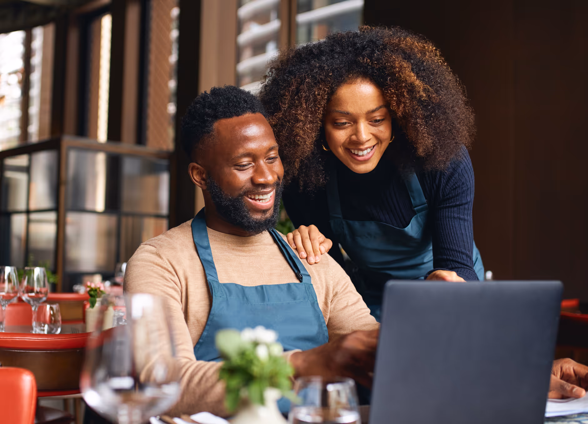 Two restaurant staff members in aprons smiling while looking at a laptop in a dining area.