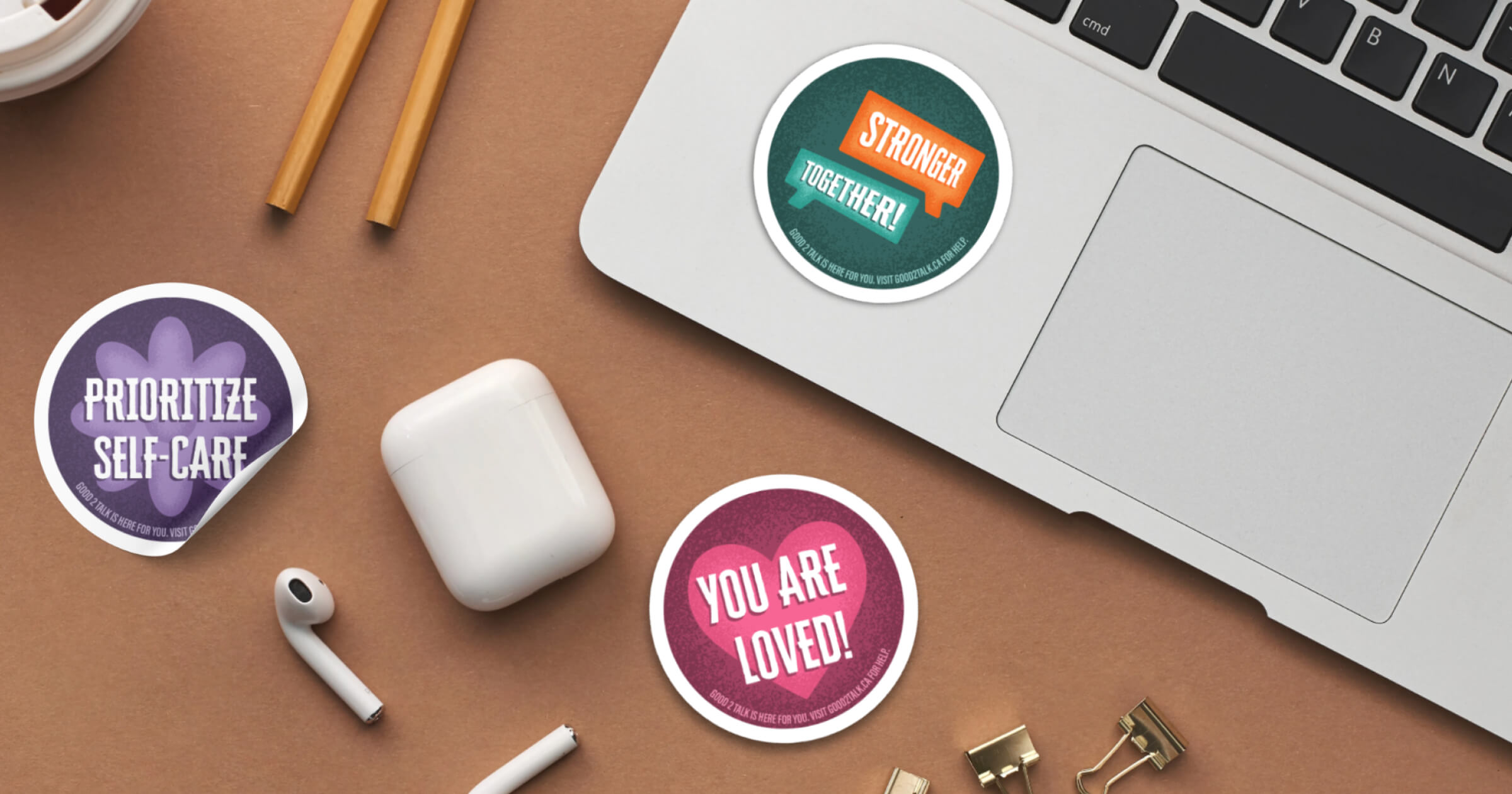 Three motivational stickers on a brown surface near a laptop and wireless earbuds, reading 'Prioritize Self-Care,' 'Stronger Together!' and 'You Are Loved!'