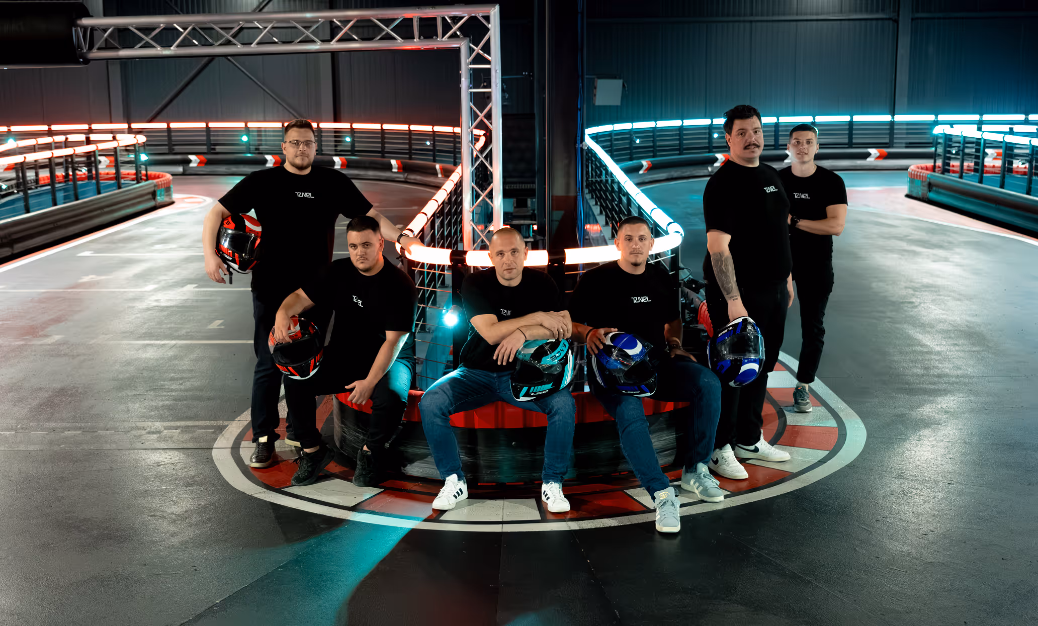 Six men in black shirts holding helmets posing at a neon-lit indoor go-kart track corner.