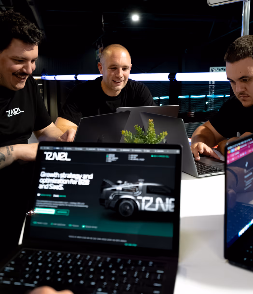 Three men working on laptops around a table with a small plant in a dark, modern office.