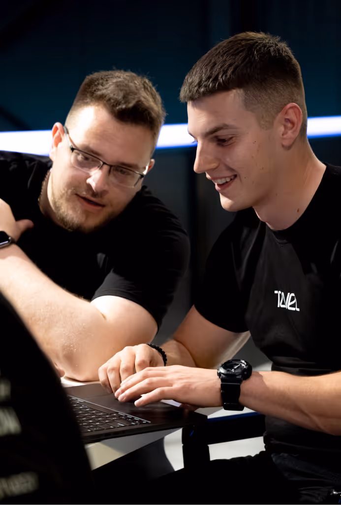 Two men in black shirts smiling and working on a laptop together indoors.