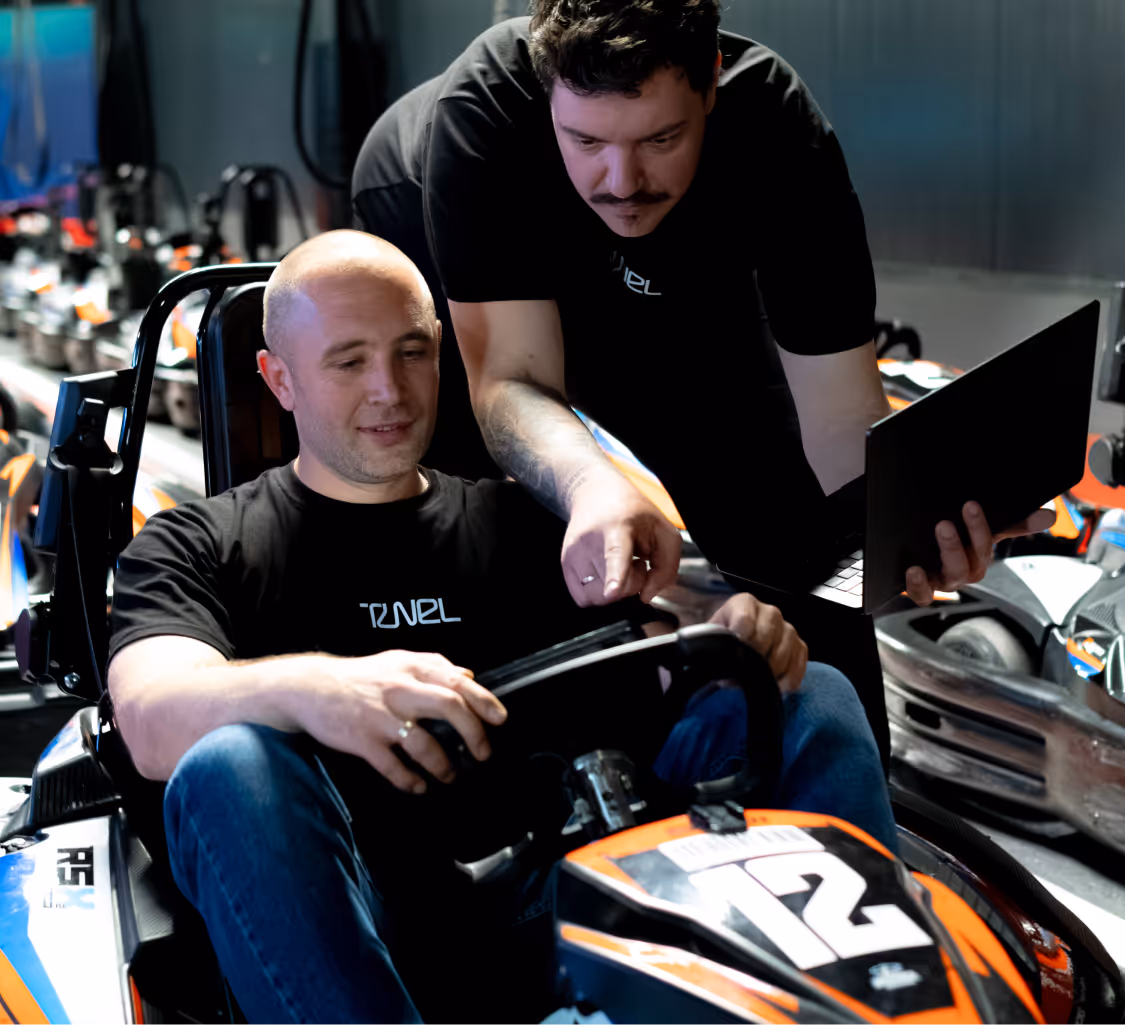 Two men in black shirts interacting over a go-kart numbered 12, one holding a laptop.