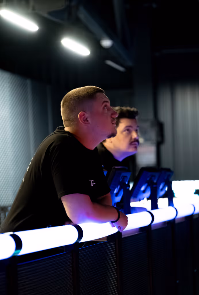 Two men leaning on a glowing railing in a dimly lit indoor space, looking upward thoughtfully.