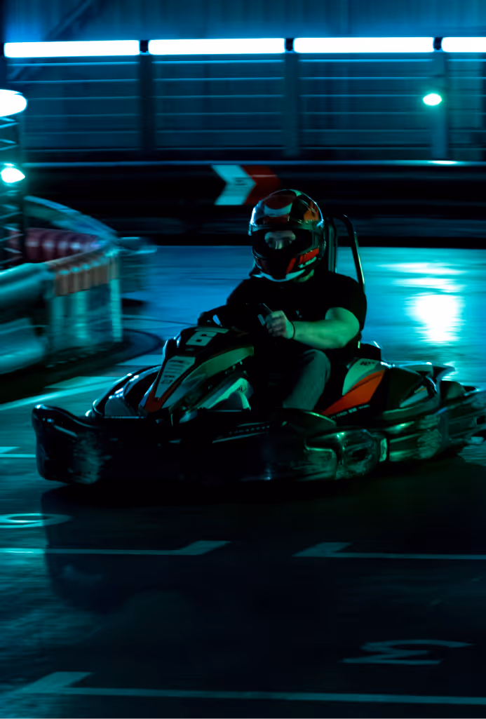 Person wearing helmet driving a go-kart on an indoor track with blue lighting.