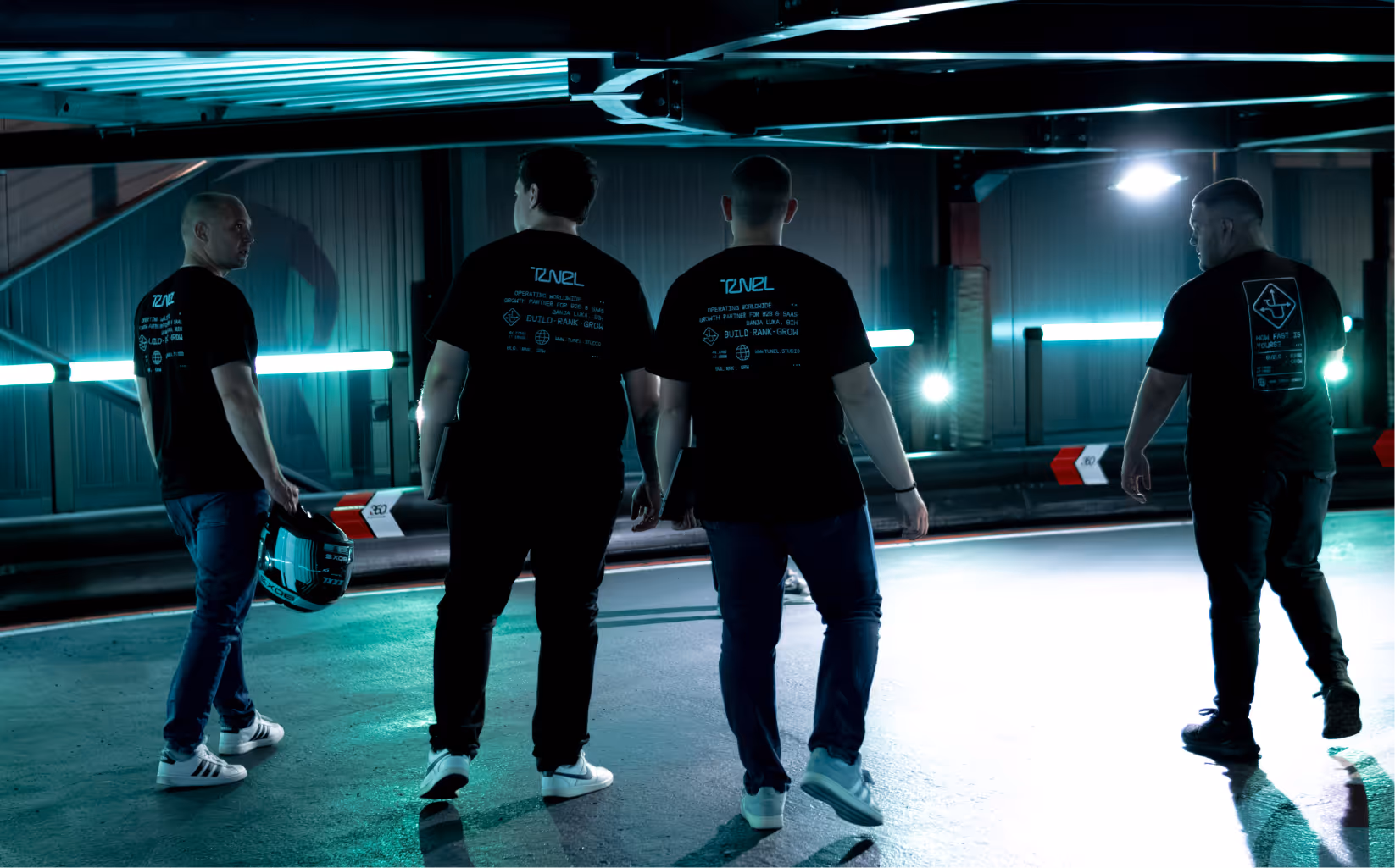 Four men wearing black T-shirts walk in an illuminated urban tunnel, one holding a motorcycle helmet.