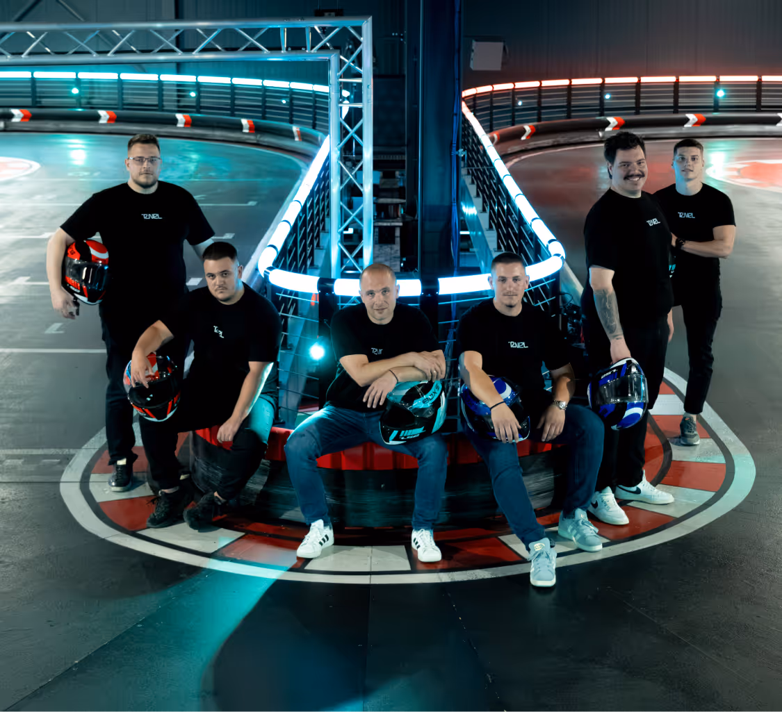 Six men in black shirts holding helmets pose at an indoor go-kart track corner with neon lighting.