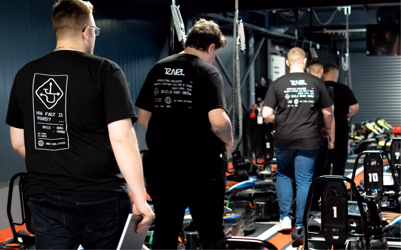Group of men in black Tunel t-shirts walk past numbered go-karts in an indoor karting area.