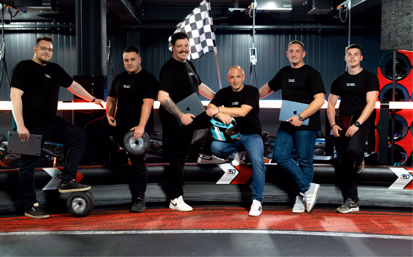 Six men in matching black shirts pose with laptops and racing gear on a karting track indoors.