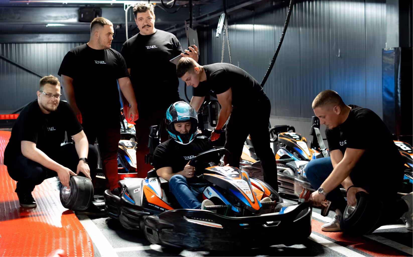 Team in black shirts preparing a go-kart racer seated with helmet inside a kart garage.