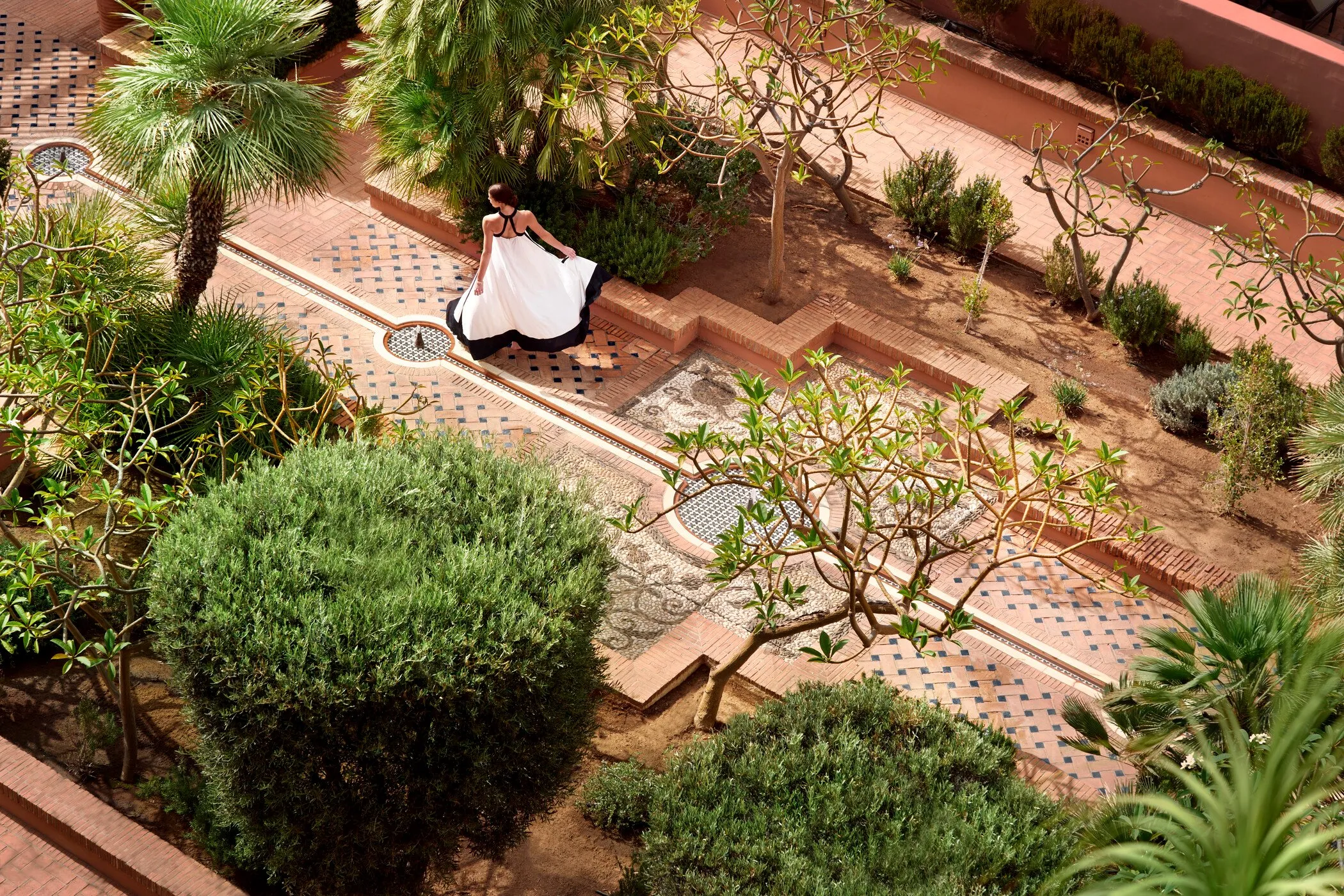 Woman in a flowing white and black dress walking on a patterned brick path surrounded by lush green plants and small trees in a garden.