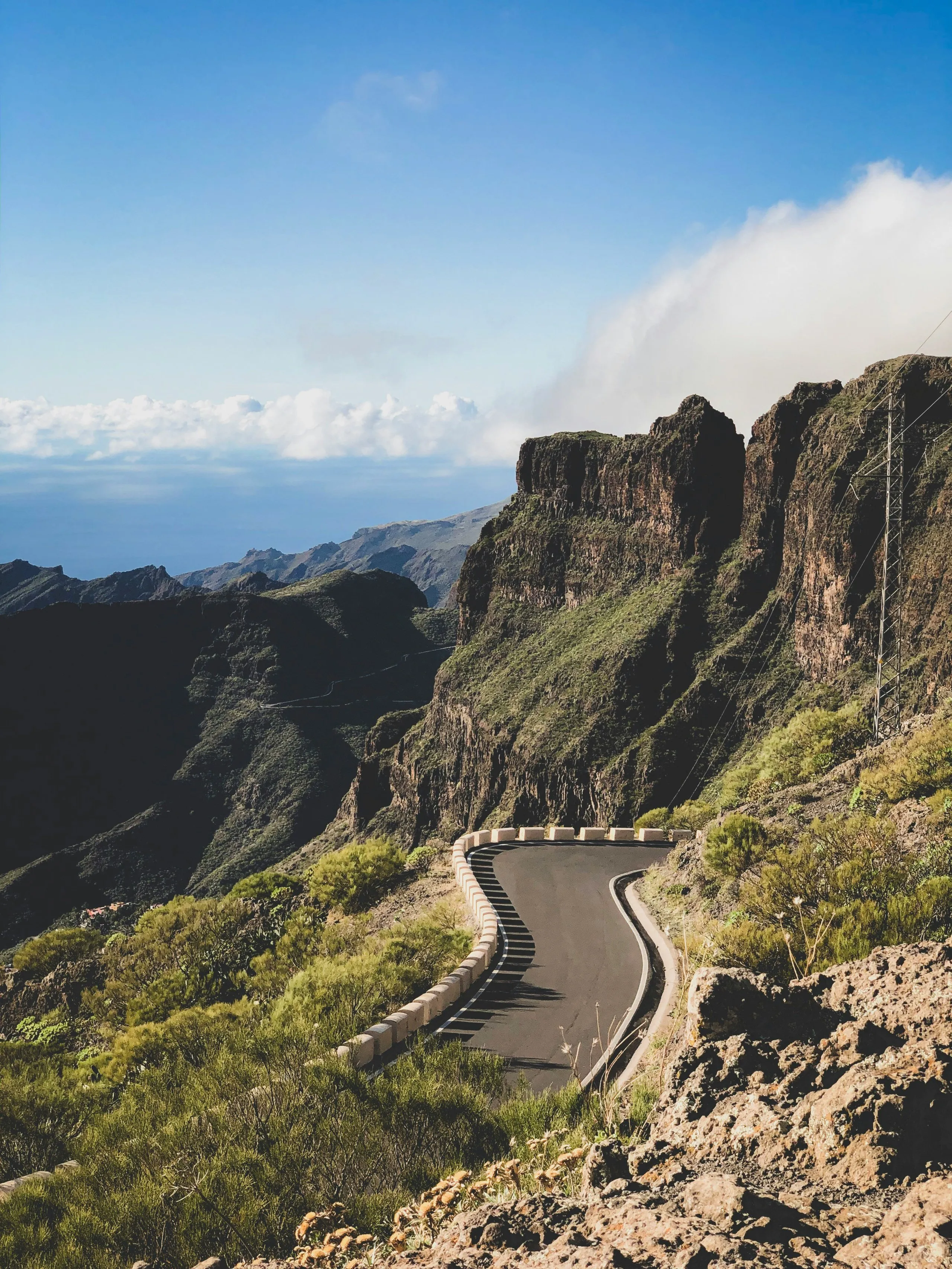 Winding mountain road lined with stone barriers surrounded by green vegetation and rocky cliffs under a blue sky.