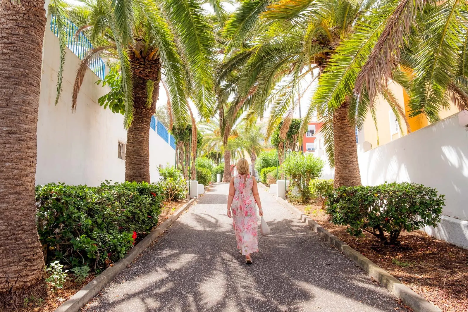 Woman in a pink floral dress walking down a palm tree-lined pathway between white walls and green bushes.