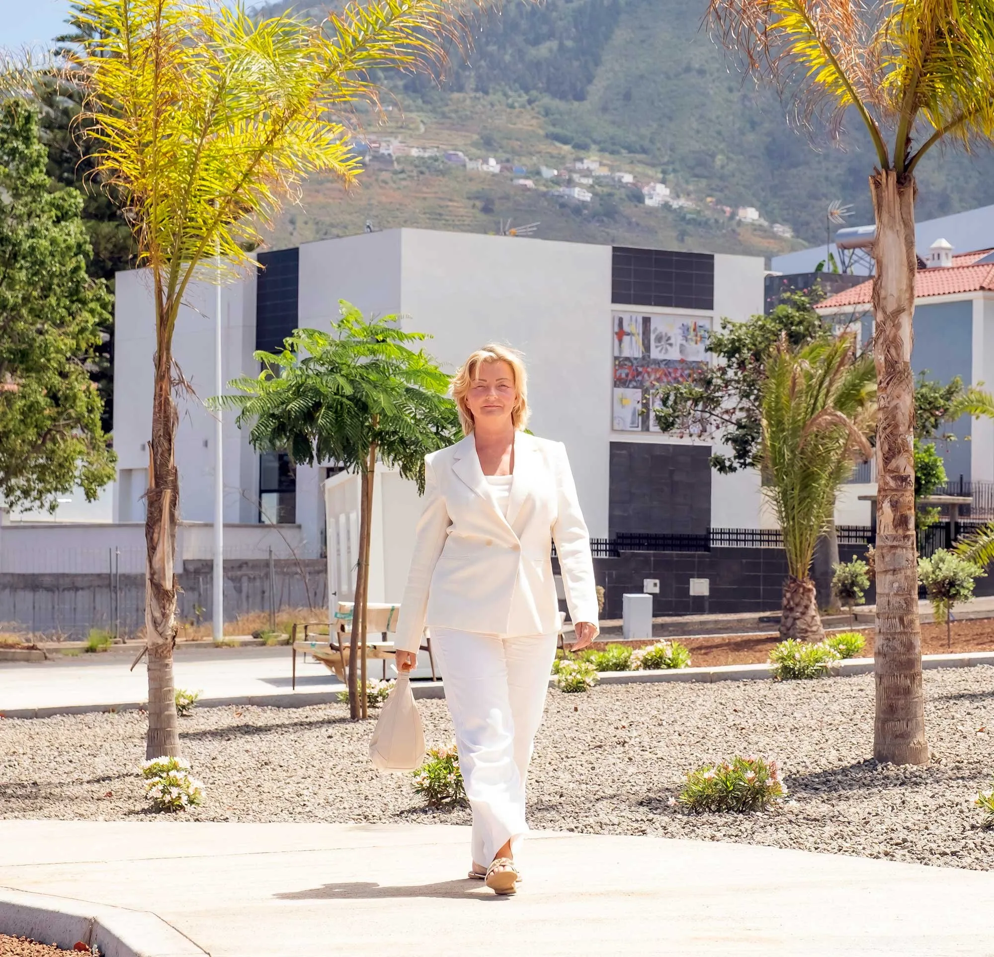Woman in a white suit walking confidently on a sunny pathway lined with palm trees and modern buildings in the background.