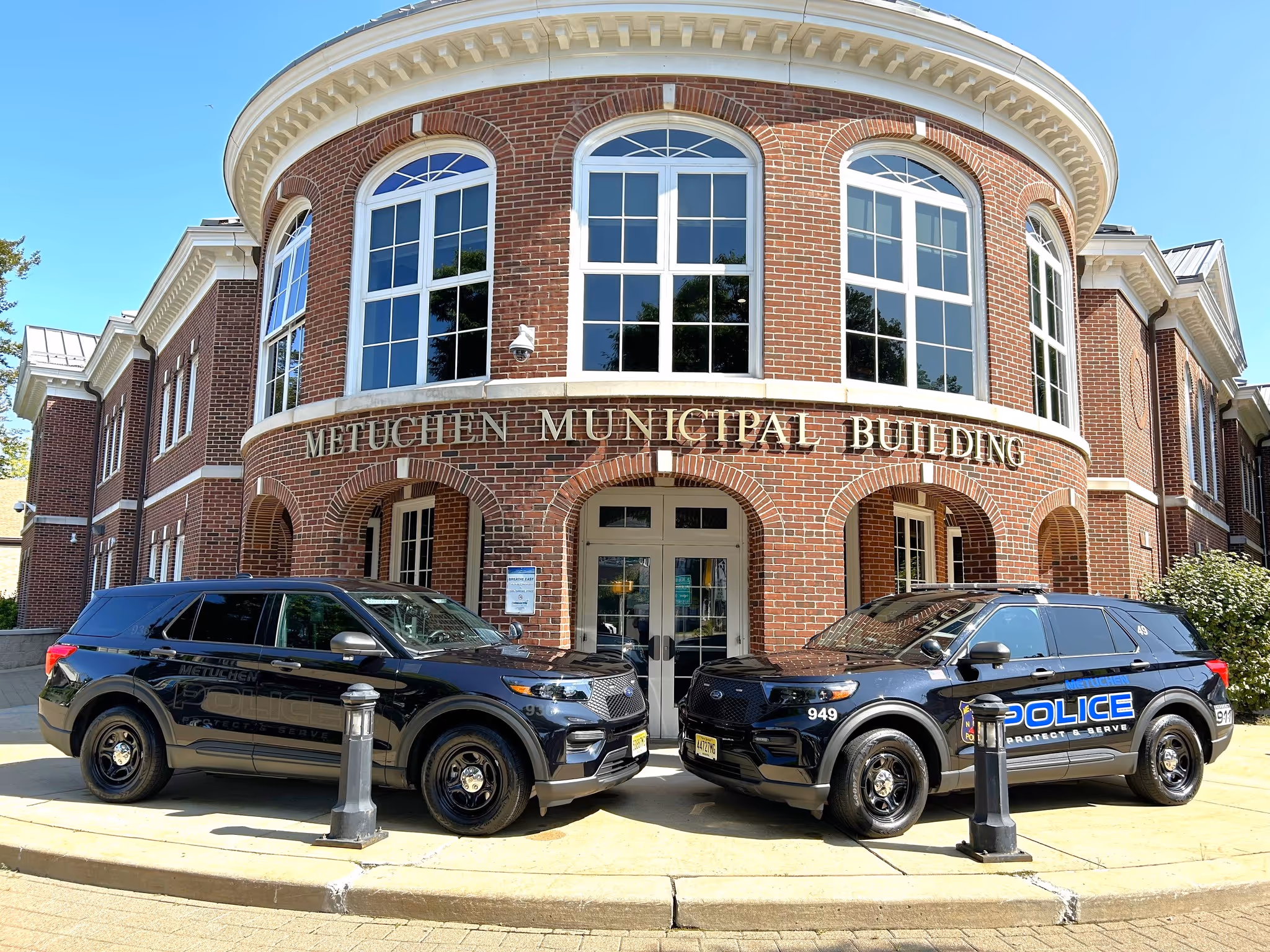 Two black police SUVs parked in front of the red brick Metuchen Municipal Building with large arched windows.