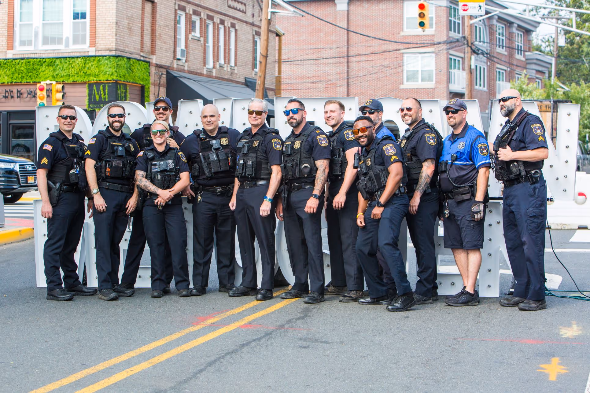 Group of uniformed police officers posing and smiling together on a city street in front of large white letters.