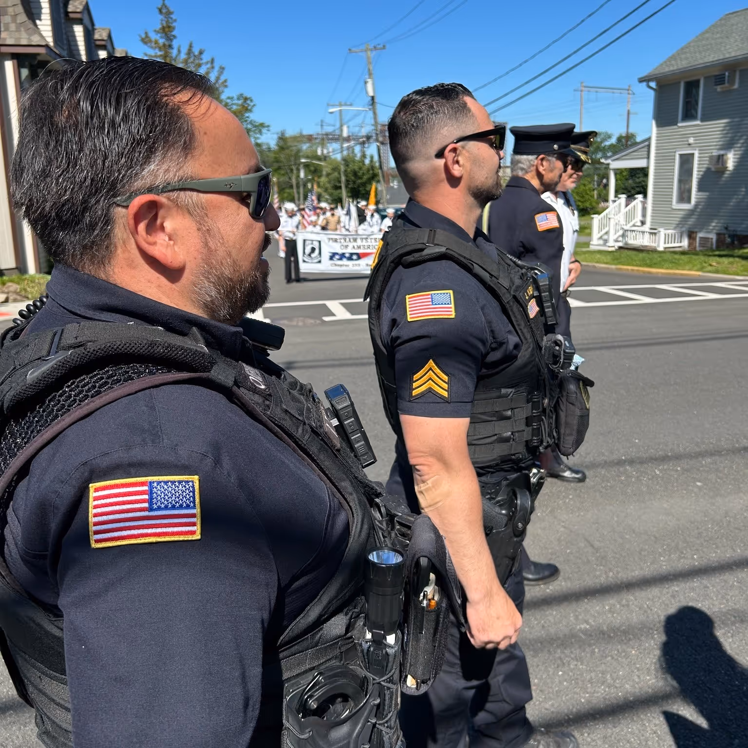 Four uniformed officers, two police and two in dress uniforms, standing in a line on a street during a daytime parade.