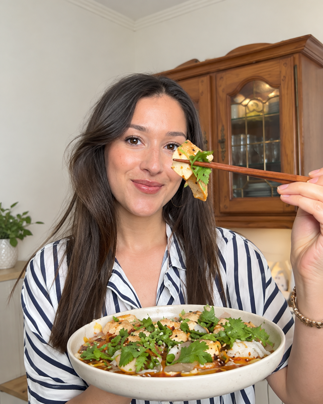 A woman with long dark hair and a striped shirt holds a bowl of food garnished with fresh herbs and uses chopsticks to lift a piece of the dish towards the camera.