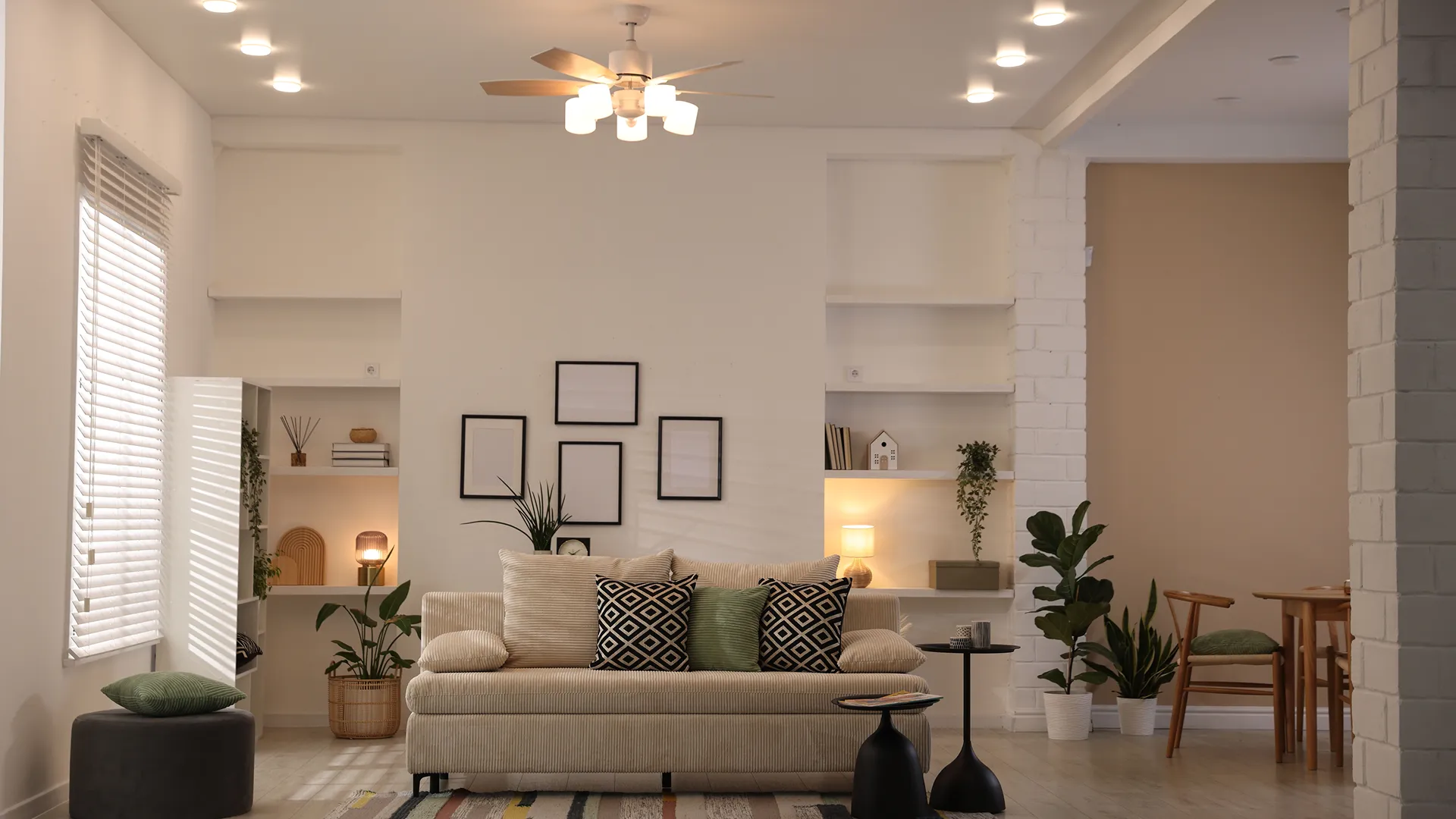 Modern living room with beige sofa, patterned and green cushions, black side tables, indoor plants, wall shelves, and neutral decor.