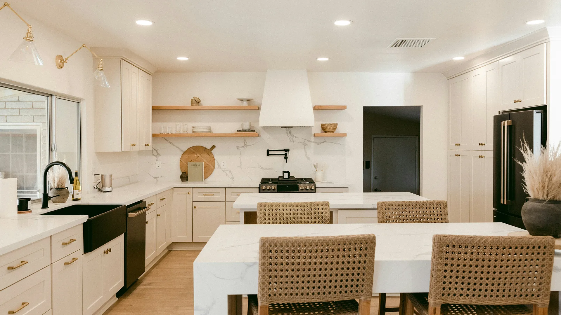 Modern kitchen with white cabinets, marble countertops, black farmhouse sink, woven chairs, and open wooden shelves.