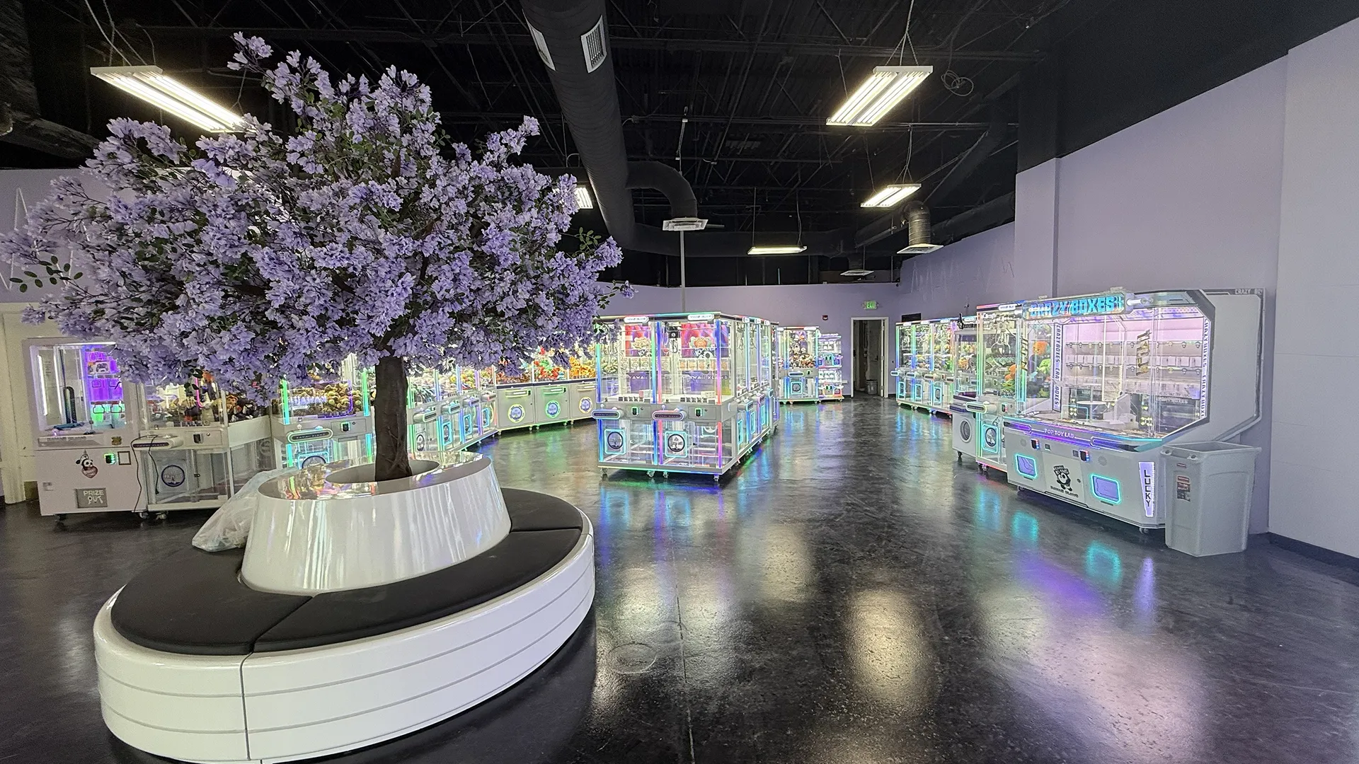 Interior of an arcade with a large purple flowering tree surrounded by circular white seating and multiple claw machines with colorful neon lights.