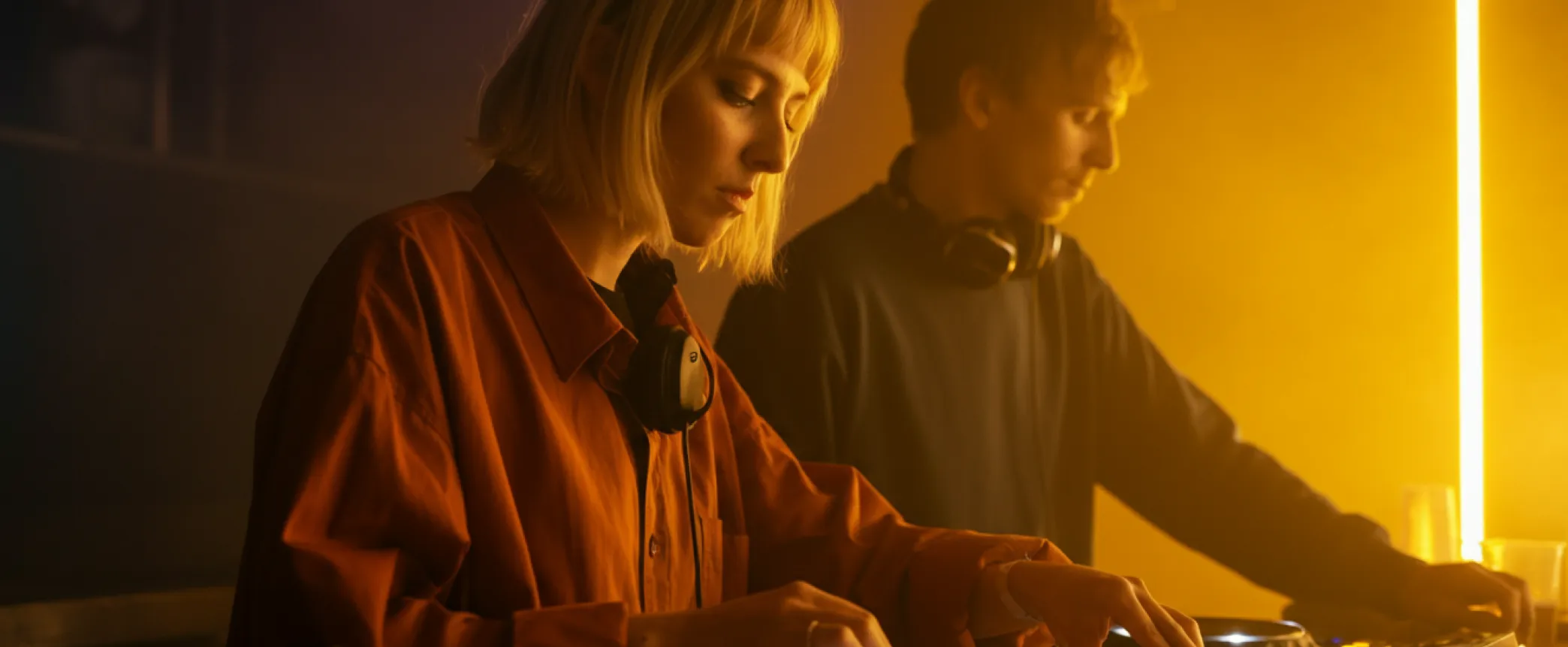 Two DJs focused on mixing music with headphones around their necks in a dimly lit environment with warm lighting.