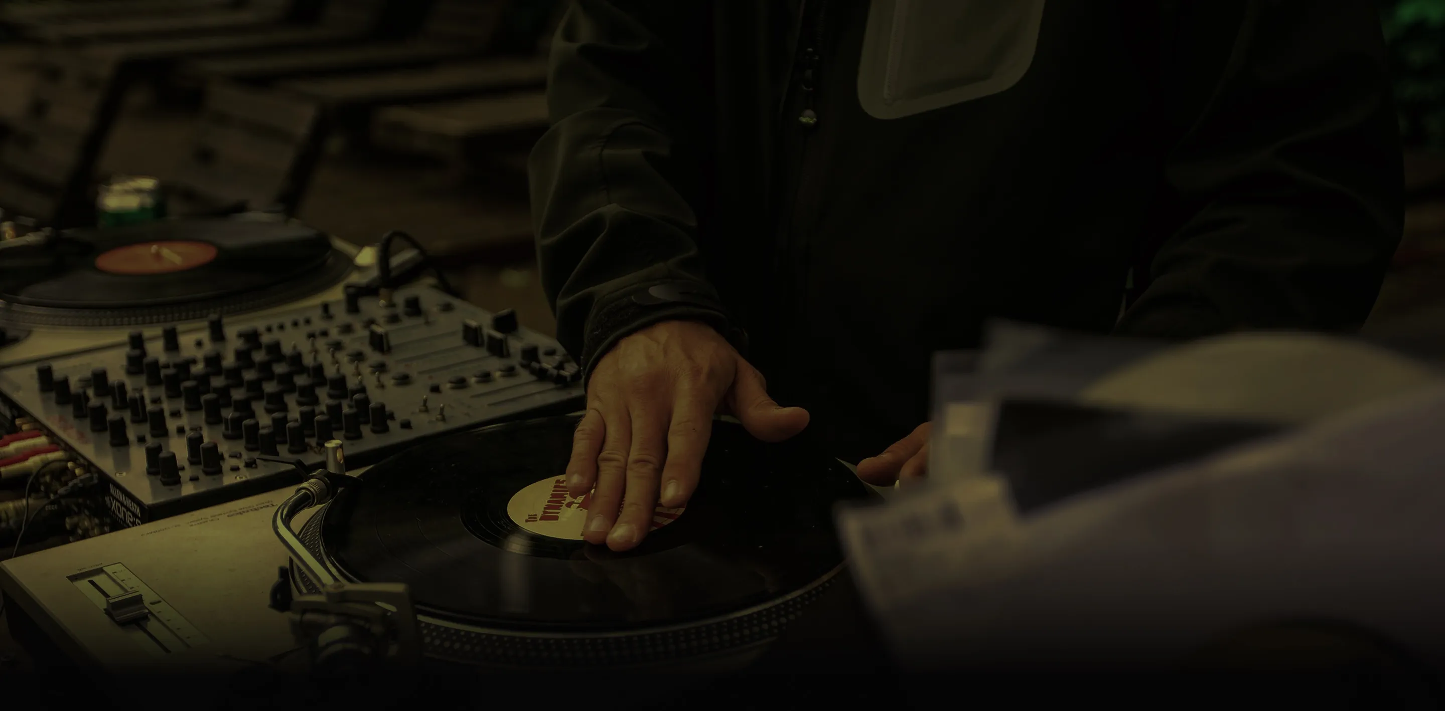 DJ's hand on a vinyl record on a turntable with a mixer in the background.