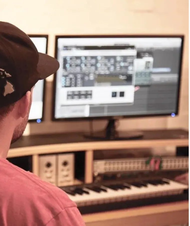 Person wearing a black cap and pink shirt working on music production software on a computer with a keyboard synthesizer in front of them.