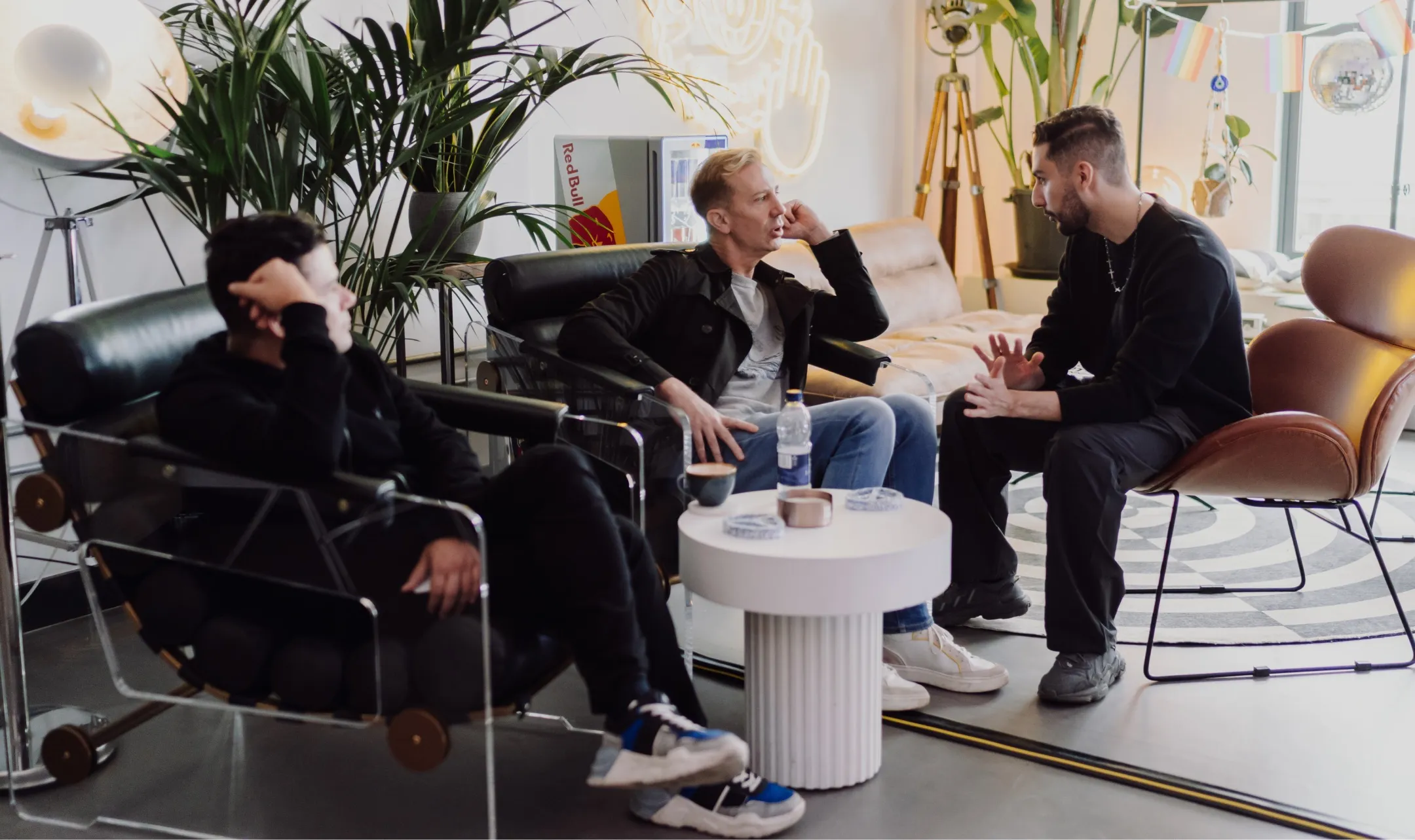 Three men engaged in conversation sitting in a modern lounge area with plants and soft lighting.