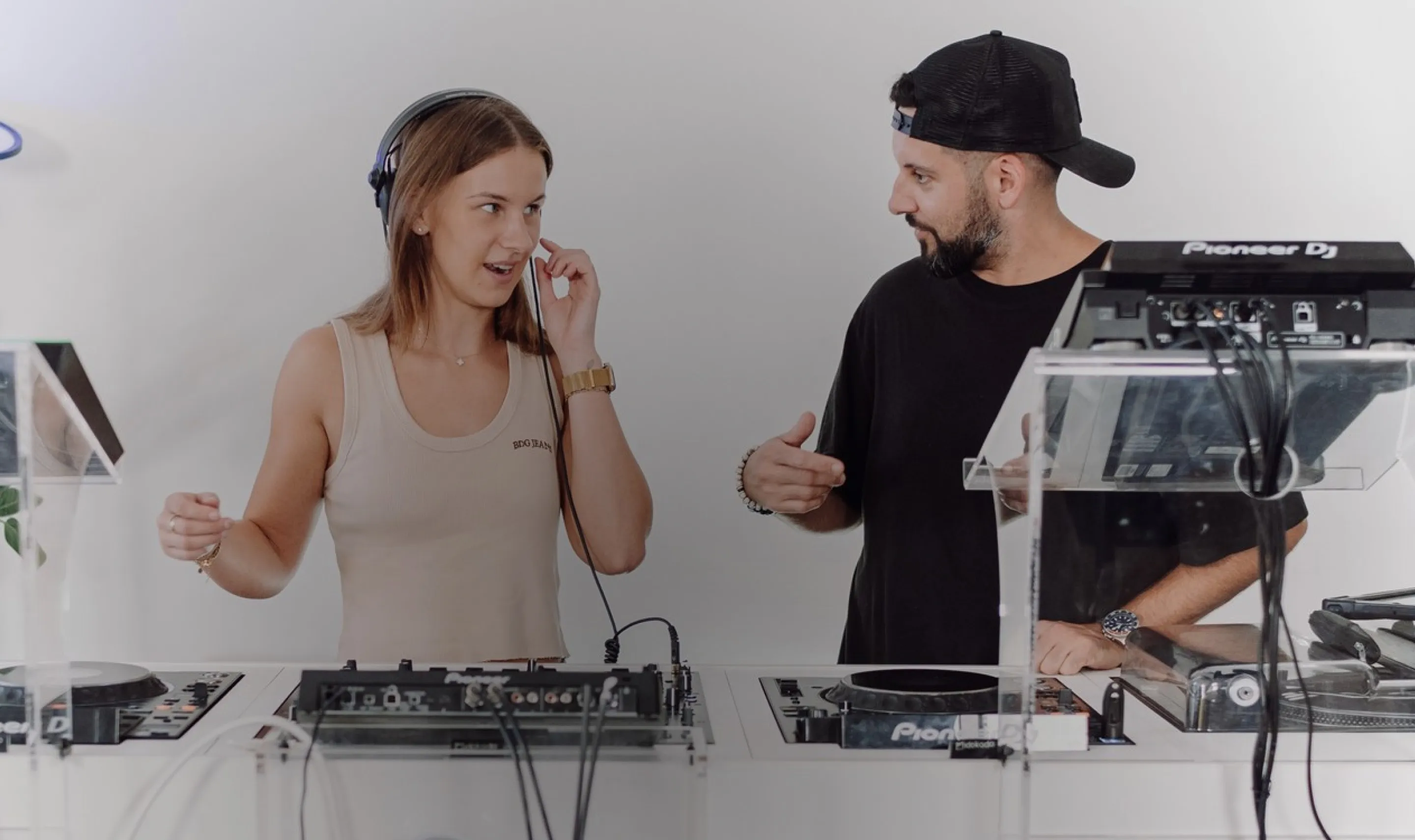 Woman with headphones adjusting DJ equipment while man in black shirt and cap gestures beside her.