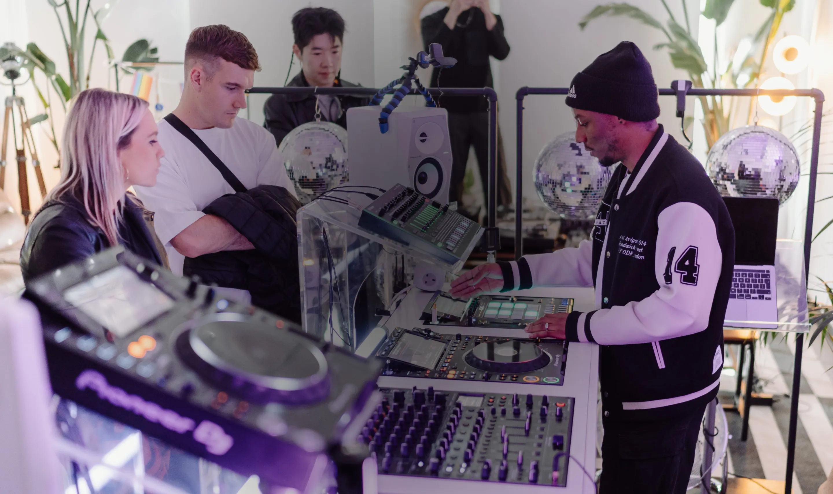 DJ performing on mixer and turntables while two people watch attentively in a modern, plant-decorated room with disco balls.