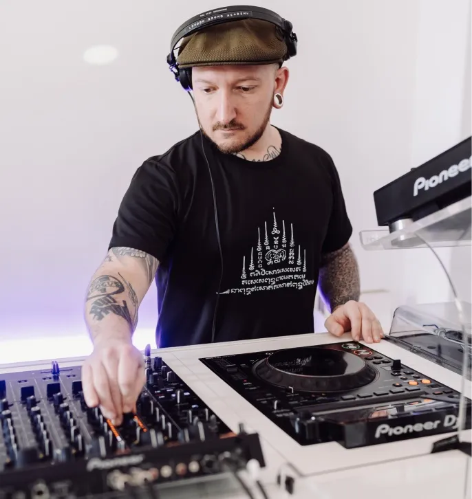 Tattooed DJ wearing a brown cap and headphones adjusting controls on a Pioneer DJ mixer.