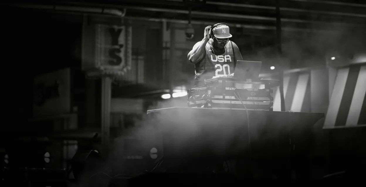 DJ wearing a USA basketball jersey and a cap mixing music on a laptop at a smoky indoor venue.