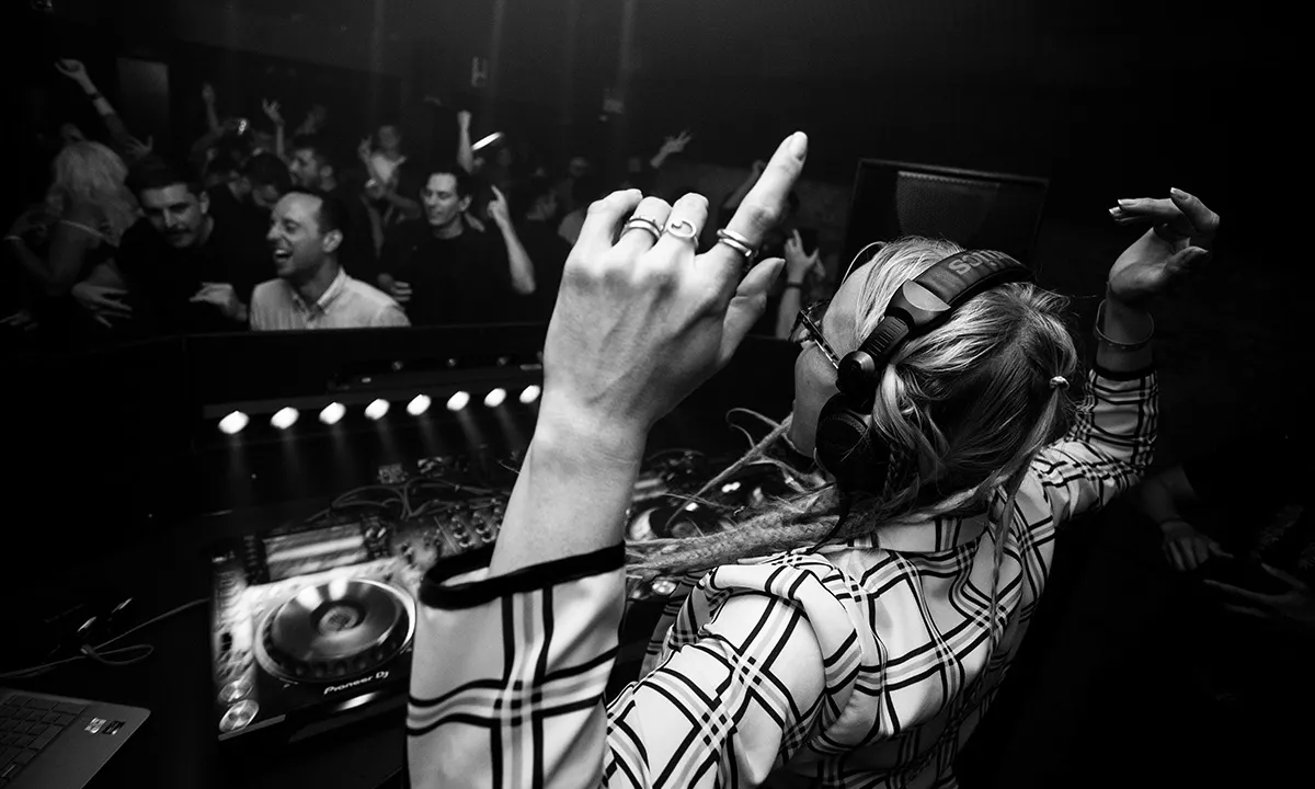 A female DJ raises her hands in the DJ booth while she drops a banger